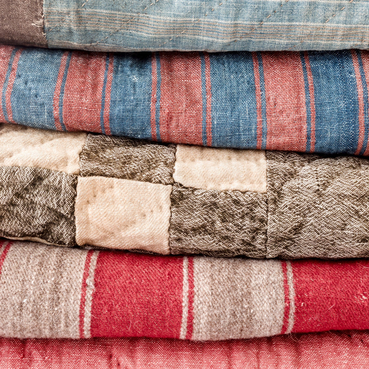 Close-up of a stack of folded antique linsey-woolsey textiles showing faded indigo blue, brick red, cream, tan, and brown stripes and checks, with visible hand stitching, irregular patchwork, softened fibers, and age-worn woven surfaces.