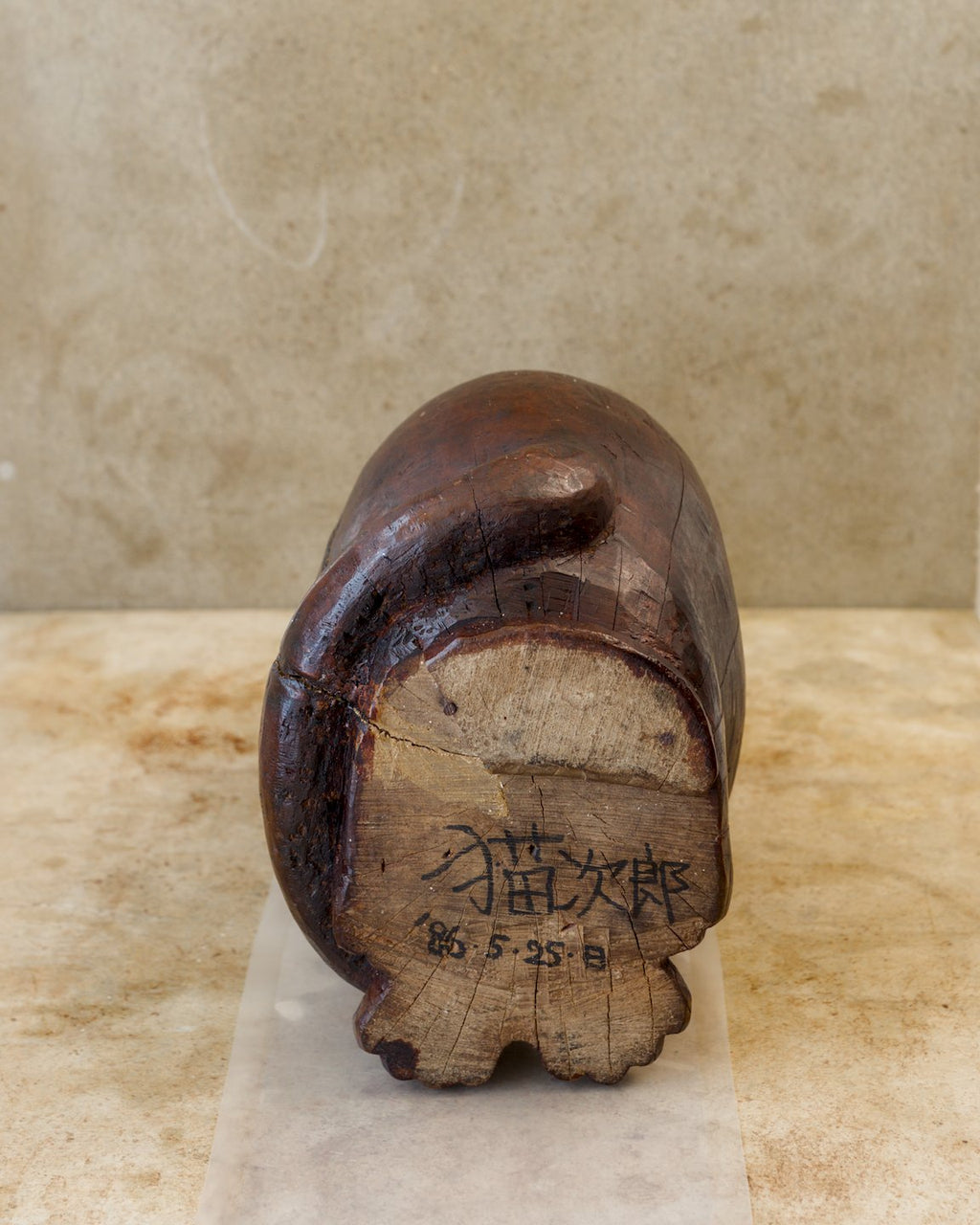 Underside of a wooden cat sculpture showing a pale, unpainted wood base with the handwritten Japanese name 猫次郎 (Nekojirō) and date 86.5.25 in black ink. The dark carved tail curves around the edge.
