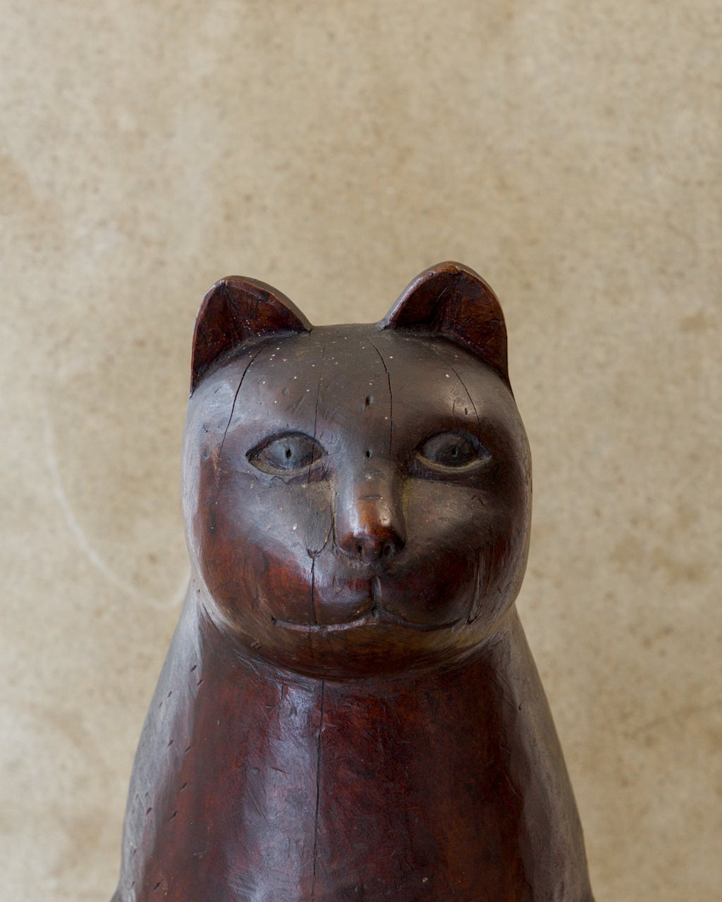 Close-up of the face and chest of a reddish-brown carved wooden cat, showing pointed ears, wide almond-shaped eyes, a small nose, a gently curved mouth, and fine lines in the wood.