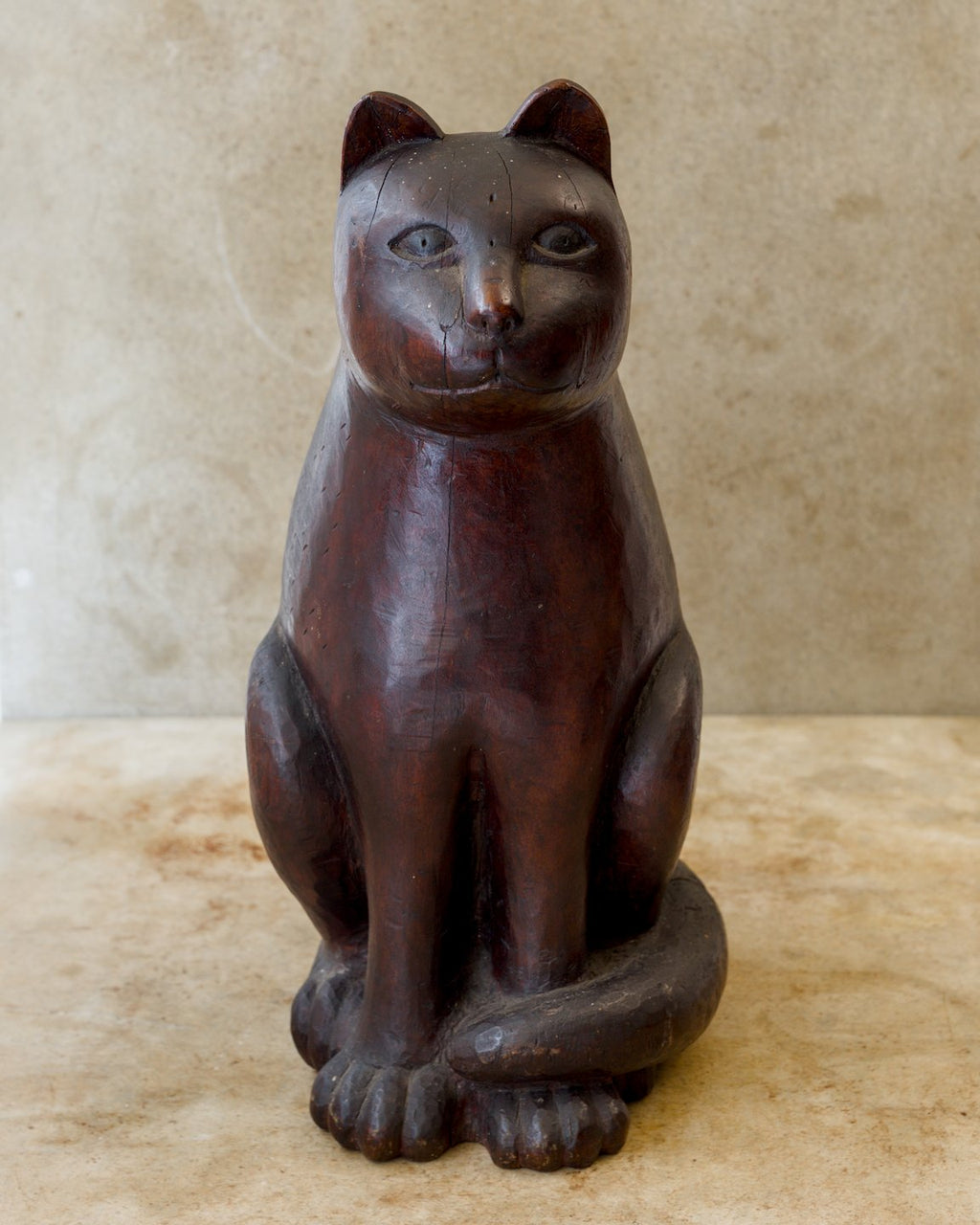 Front view of a seated Japanese wooden cat sculpture with a reddish-brown finish, upright ears, wide-set eyes, broad front paws, and a thick tail curled across its feet.