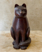 Front view of a seated Japanese wooden cat sculpture with a reddish-brown finish, upright ears, wide-set eyes, broad front paws, and a thick tail curled across its feet.
