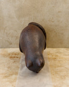 Overhead view of a dark reddish-brown carved wooden cat sculpture, showing its broad, rounded back, two pointed ears, and thick curled tail. Fine carving marks, small pale specks, and visible separations in the wood follow the contours of the body.