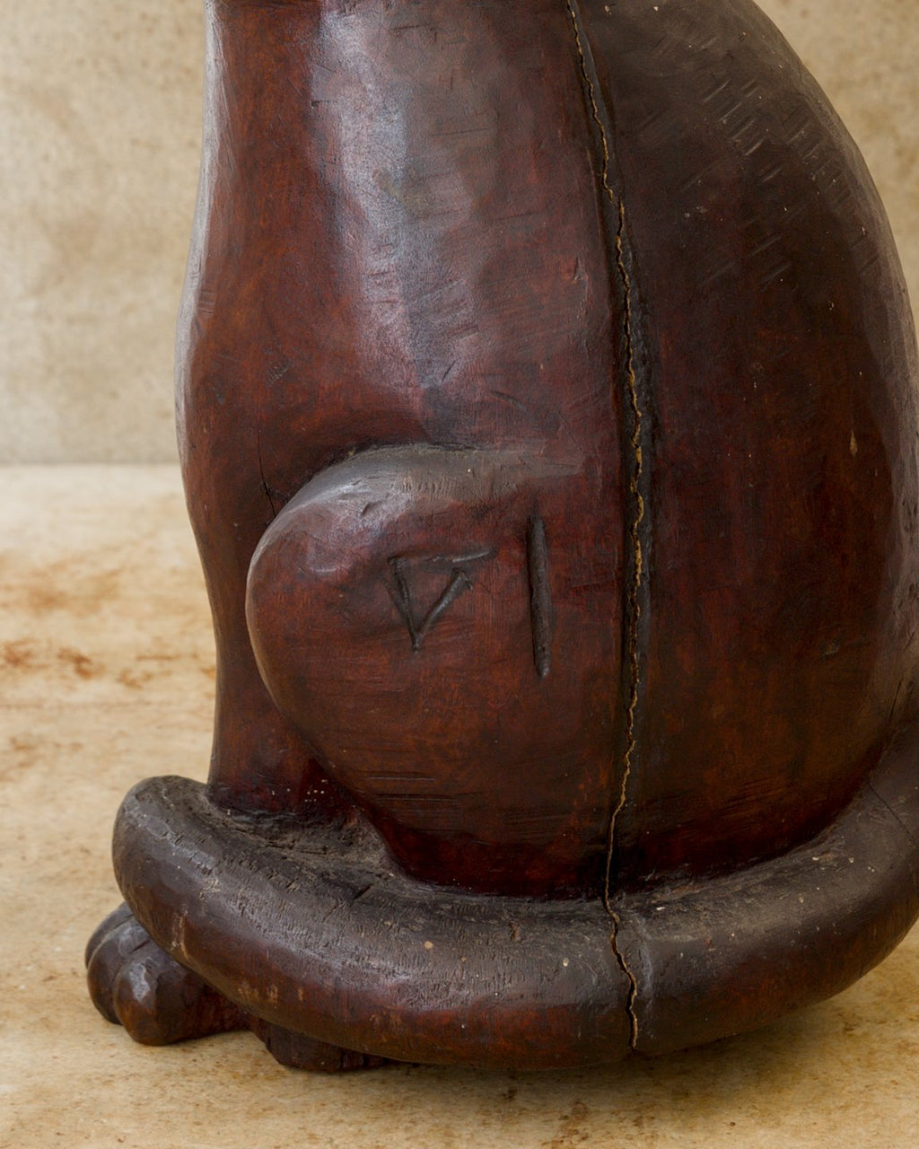 Close-up of a reddish-brown carved wooden cat sculpture showing an angular incised mark on the rounded haunch, a long separation in the wood beside it, and a thick tail curling around the base.