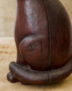 Close-up of a reddish-brown carved wooden cat sculpture showing an angular incised mark on the rounded haunch, a long separation in the wood beside it, and a thick tail curling around the base.