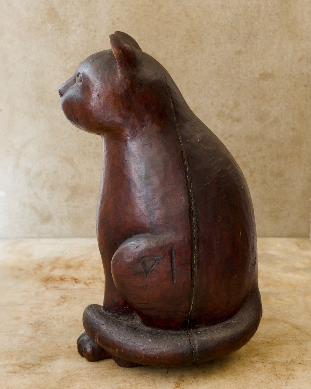 Left-facing profile of a reddish-brown carved wooden cat, with pointed ears, a rounded muzzle, a small carved symbol on its haunch, and a thick tail circling the base. A visible separation follows the wood grain down its side.