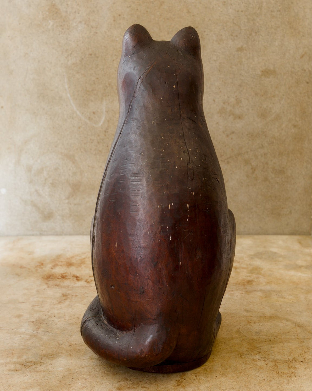 Rear view of a seated reddish-brown wooden cat sculpture, showing a tall rounded back with scattered carving marks, two pointed ears, and a thick tail wrapped around the base.