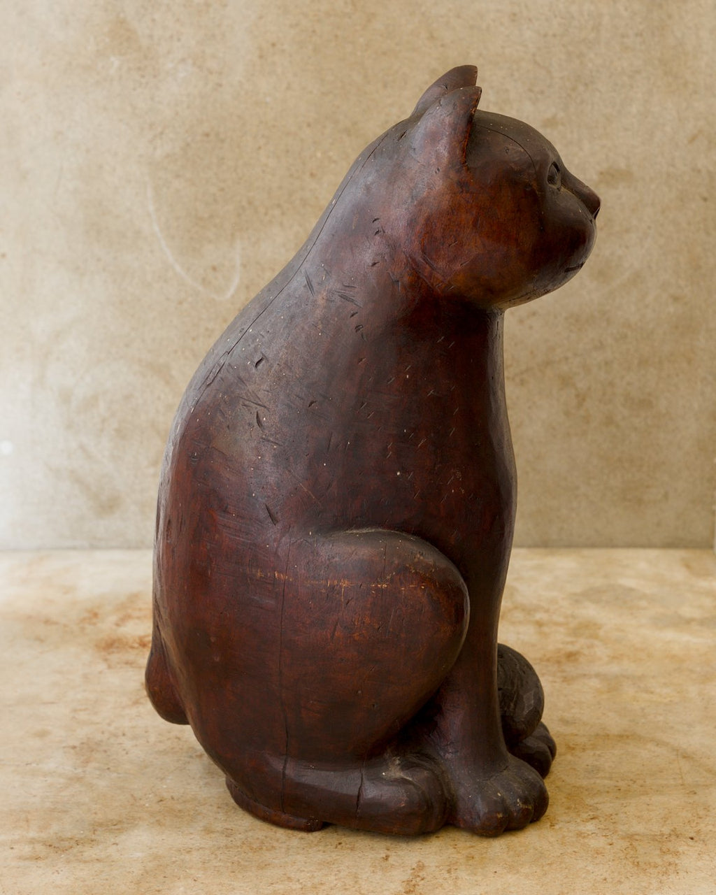 Right-facing profile of a seated wooden cat sculpture with a rounded body, upright pointed ears, thick front paws, and a tail curled around the base. Fine tool marks are visible in its dark reddish-brown surface.
