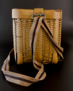 Vintage Japanese bamboo biku fishing basket viewed from its narrow side. A beige strap with a dark stripe edged in red hangs in long folds from a metal loop at the rim and trails onto the black surface below.