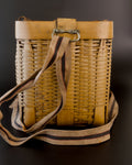 Narrow end of a vintage Japanese biku fishing basket in golden bamboo. A beige carrying strap with a dark stripe edged in red hangs from a metal loop, crosses the woven side, and curls around the base.