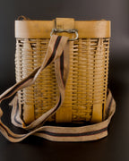 Narrow end of a vintage Japanese biku fishing basket in golden bamboo. A beige carrying strap with a dark stripe edged in red hangs from a metal loop, crosses the woven side, and curls around the base.