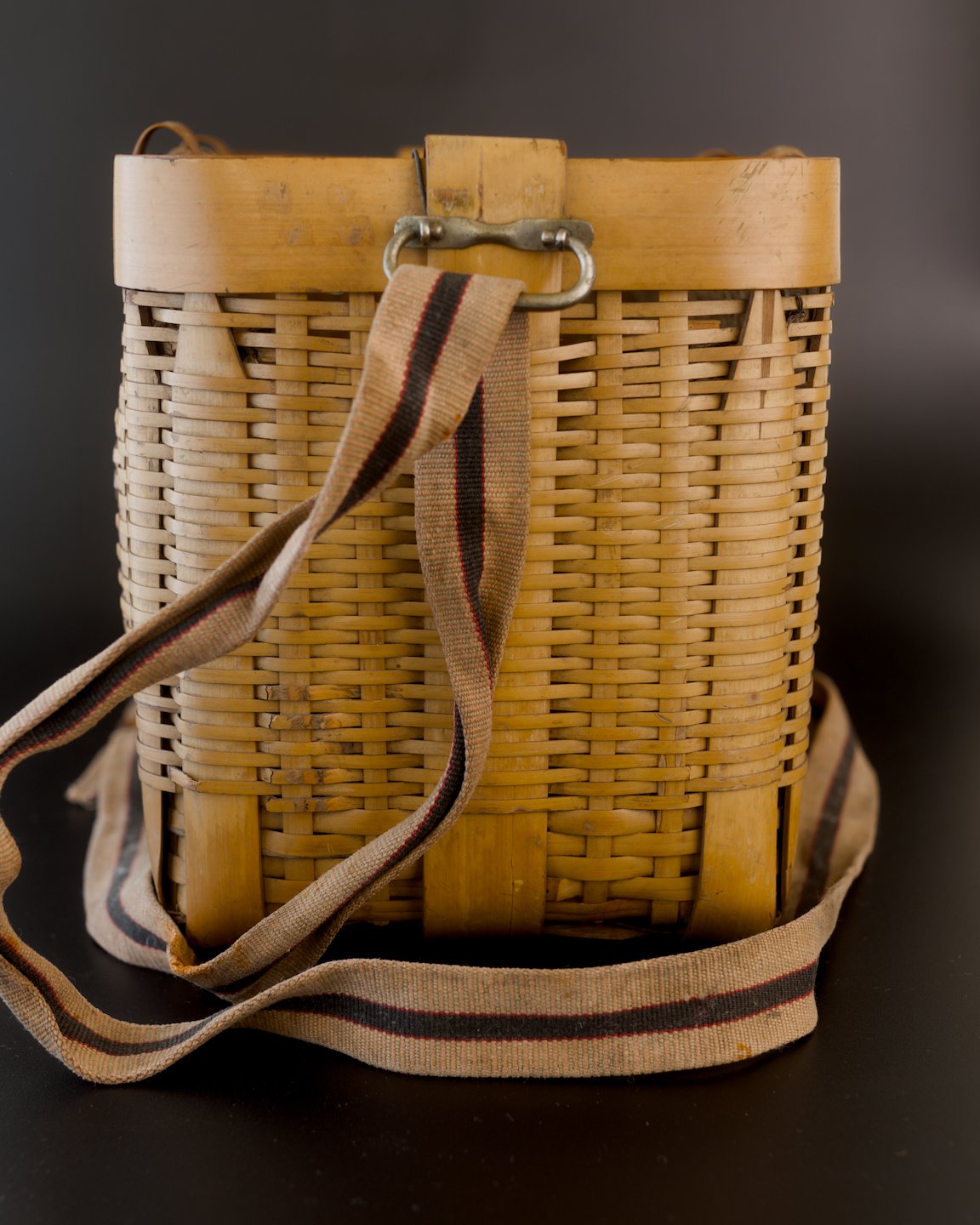 Narrow end of a vintage Japanese biku fishing basket in golden bamboo. A beige carrying strap with a dark stripe edged in red hangs from a metal loop, crosses the woven side, and curls around the base.