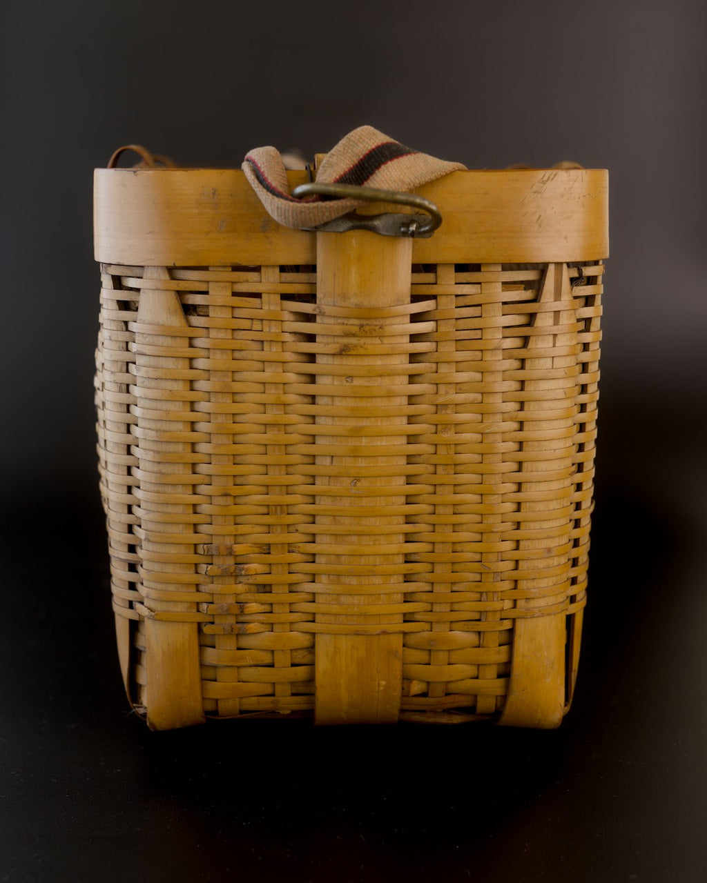 Narrow end of a vintage Japanese bamboo biku fishing basket, with the striped fabric strap folded over its broad rim. Fine horizontal bamboo strips cross wider upright ribs, with darker patches near the base.