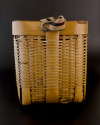 Narrow side of a vintage Japanese biku fishing basket, with its striped fabric strap folded above a metal attachment loop. The golden bamboo has a fine horizontal weave, wide upright supports, and a darker worn patch at the upper right rim.