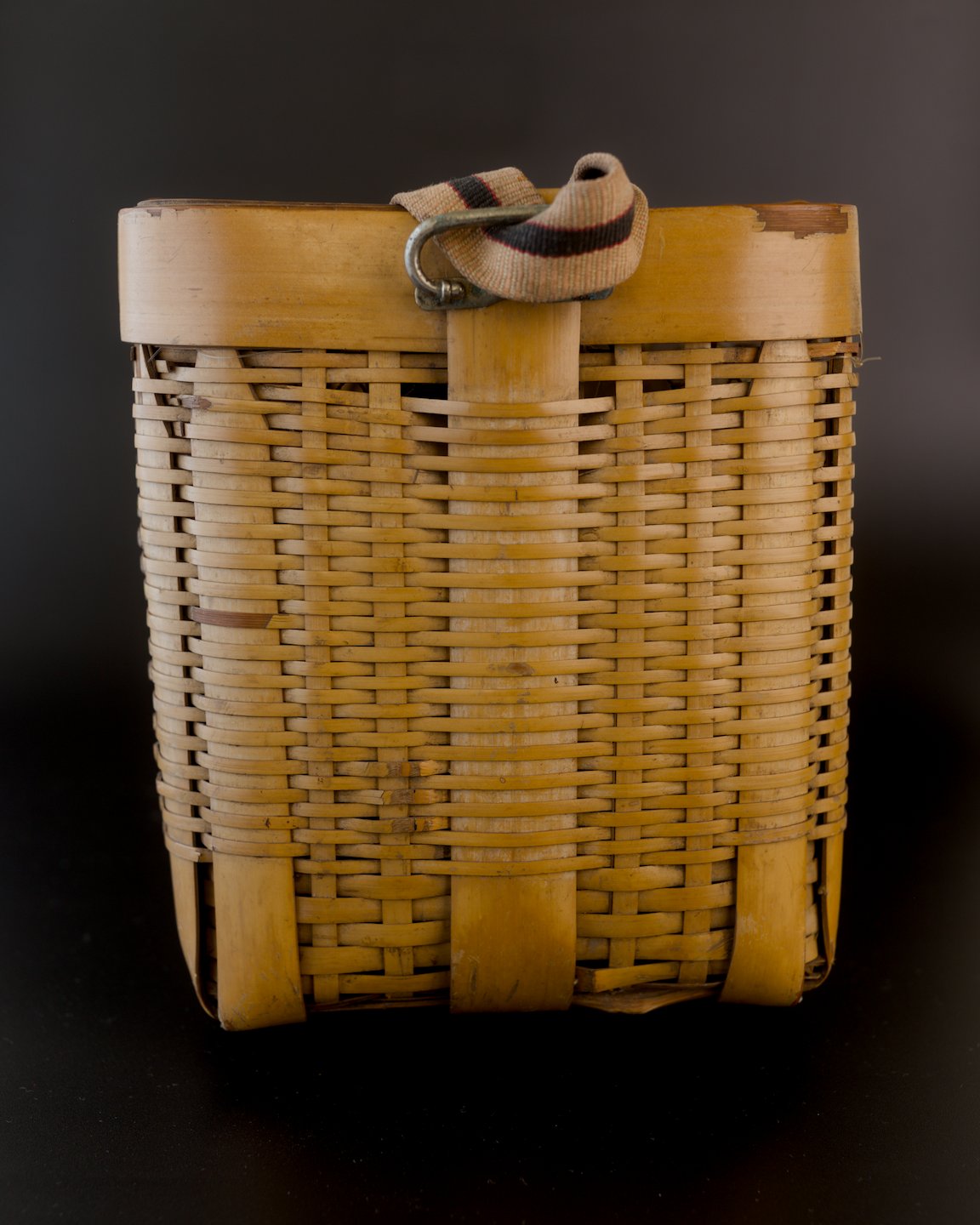 Narrow side of a vintage Japanese biku fishing basket, with its striped fabric strap folded above a metal attachment loop. The golden bamboo has a fine horizontal weave, wide upright supports, and a darker worn patch at the upper right rim.