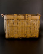 Broad side of a vintage Japanese bamboo biku fishing basket, viewed slightly from above. Two wide vertical bands cross the finely woven body, thin bindings encircle the rim, and ends of the striped strap rest at the upper corners.