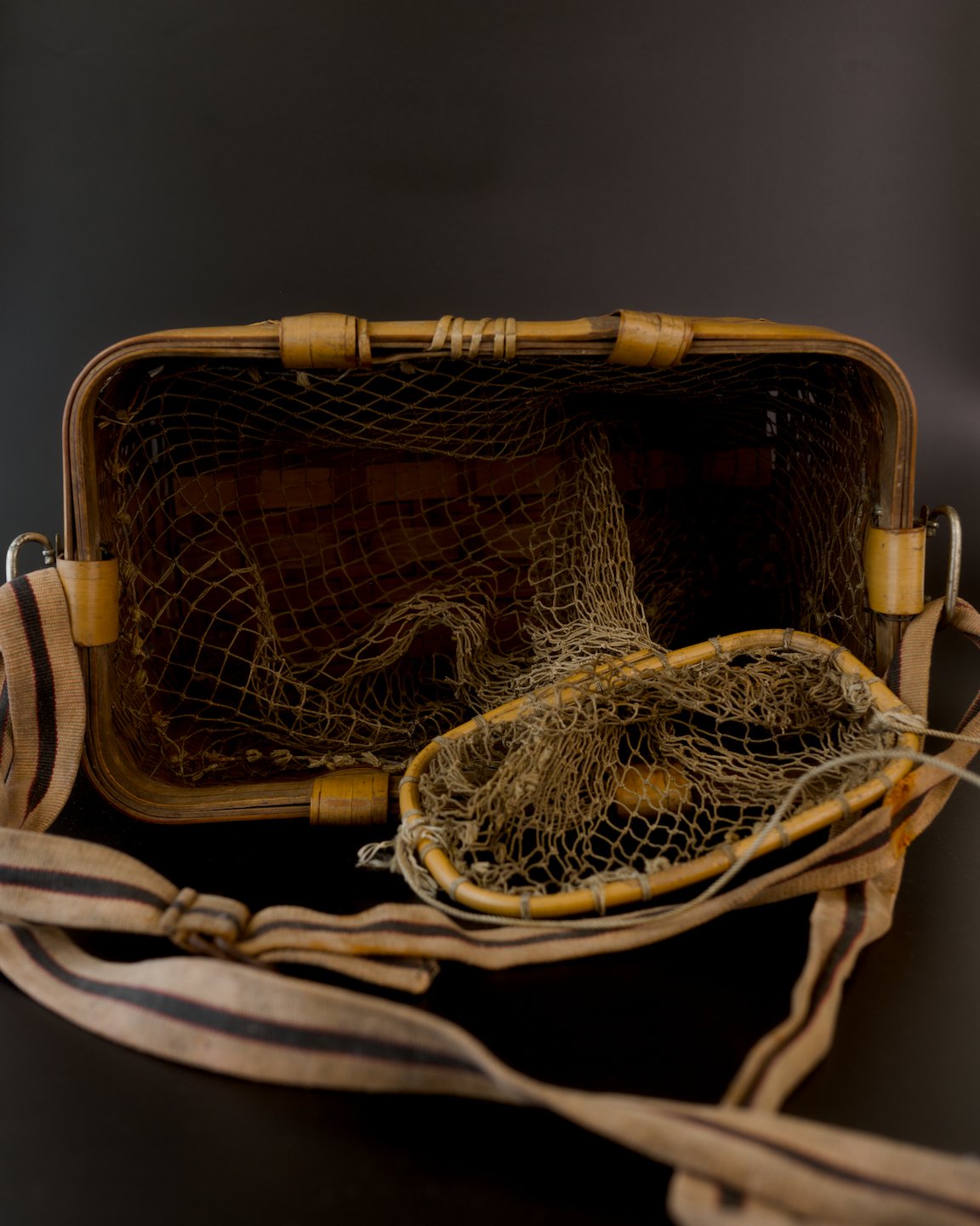 View into a vintage Japanese bamboo biku fishing basket. Brown knotted netting lines the rectangular opening, with its small bamboo-ring mouth pulled out toward the foreground. A beige, dark-striped strap and metal buckle lie across the black surface.