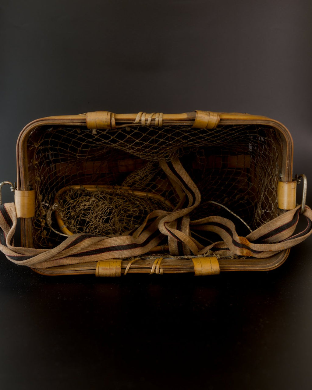 Overhead view into a vintage Japanese bamboo biku fishing basket. Brown netting spans the rounded rectangular rim; a small bamboo-ring opening sits inside at the left. The beige strap with dark, red-edged stripes coils across the interior and lower rim.