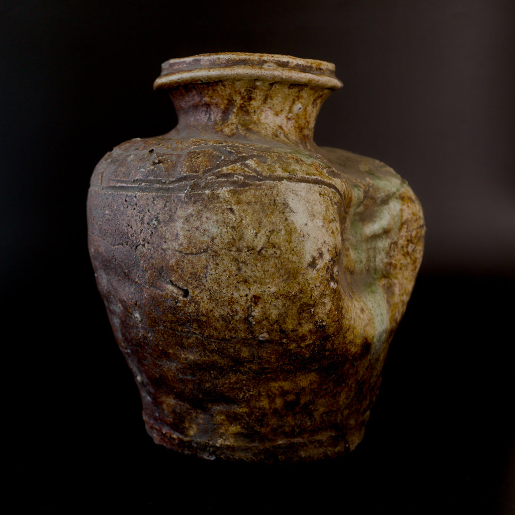 Three-quarter view of an antique Shigaraki stoneware jar, with incised zigzag decoration near the neck and a sunken side coated with patches of pale green natural ash glaze.