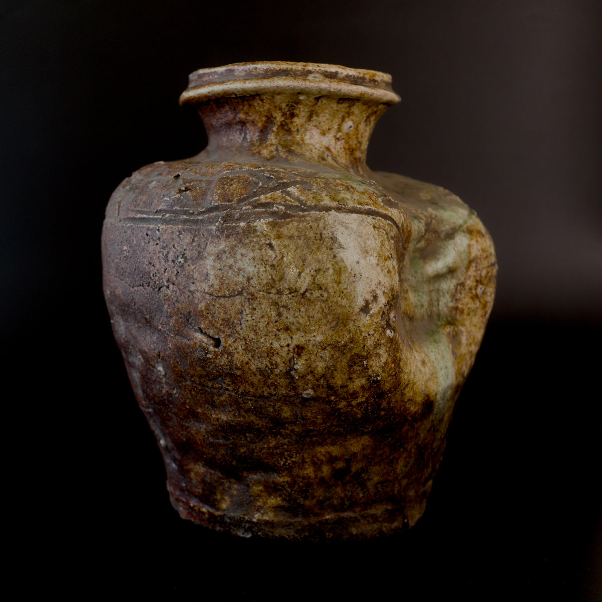 Three-quarter view of an antique Shigaraki stoneware jar, with incised zigzag decoration near the neck and a sunken side coated with patches of pale green natural ash glaze.