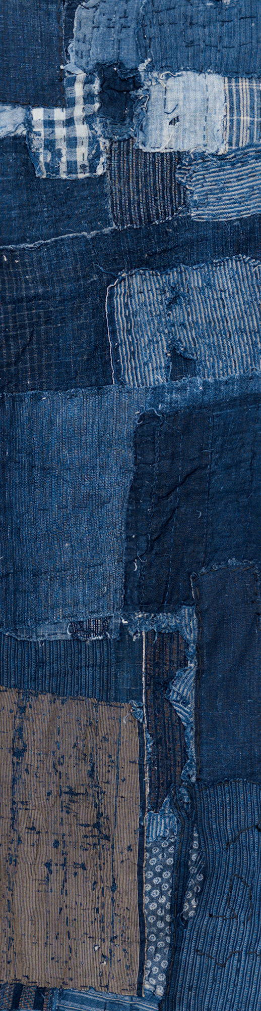 Cropped, long, narrow antique Japanese boro cotton panel displayed vertically. Rectangular patches of dark and faded indigo cotton include checked and striped fabrics near the top, larger blue sections through the center, and a worn tan patch near the lower left. Hand stitching, and loose threads.