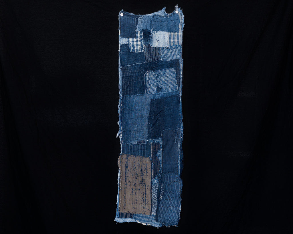 Long, narrow antique Japanese boro cotton panel displayed vertically against a black background. Rectangular patches of dark and faded indigo cotton include checked and striped fabrics near the top, larger blue sections through the center, and a worn tan patch near the lower left. Hand stitching, loose threads, and frayed edges are visible throughout.