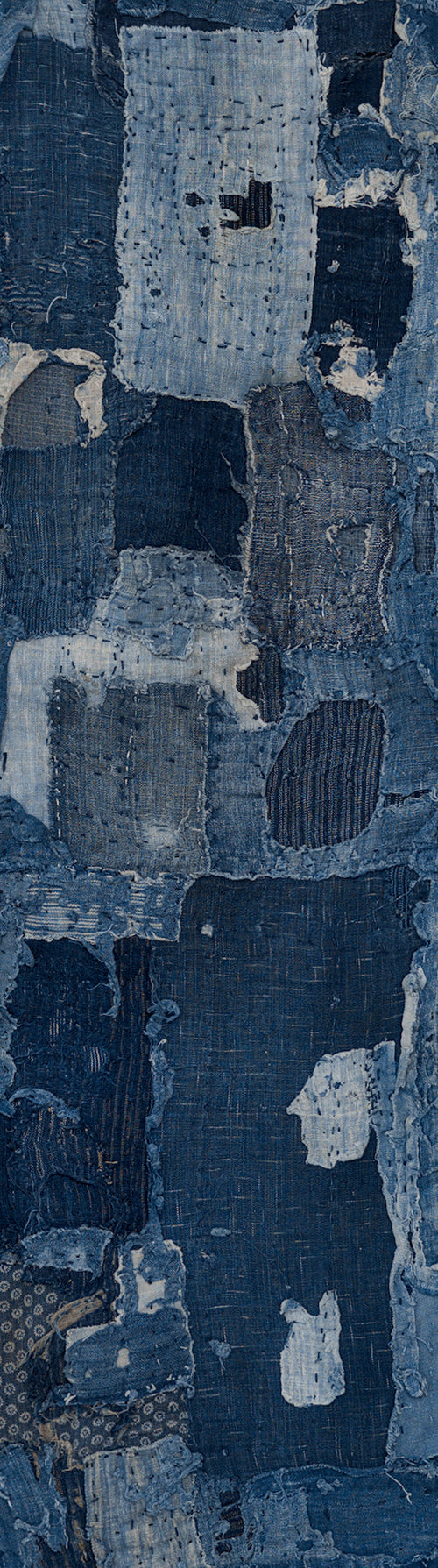 Vista ritagliata di un lungo e stretto pannello boro giapponese antico in cotone, disposto in verticale. Toppe sovrapposte in indaco scuro e sbiadito, cuciture a mano e zone consumate. Grande toppa azzurro chiaro in alto e piccoli tessuti fantasia in basso.