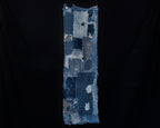 Long, narrow antique Japanese boro cotton panel displayed vertically against a black background. Overlapping patches of dark and faded indigo cotton feature extensive hand stitching, frayed edges, and worn-through areas. A large pale blue patch appears near the top, with smaller patterned cotton patches near the bottom.