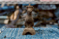 A small antique Japanese clay Inari fox sits upright, facing forward, with pointed ears, a long neck, a rounded chest, and narrow front legs. Its rough charcoal-brown surface extends to a pedestal with a large arched opening, resting on frayed blue woven cloth.