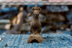 A small antique Japanese clay Inari fox sits upright, facing forward, with pointed ears, a long neck, a rounded chest, and narrow front legs. Its rough charcoal-brown surface extends to a pedestal with a large arched opening, resting on frayed blue woven cloth.