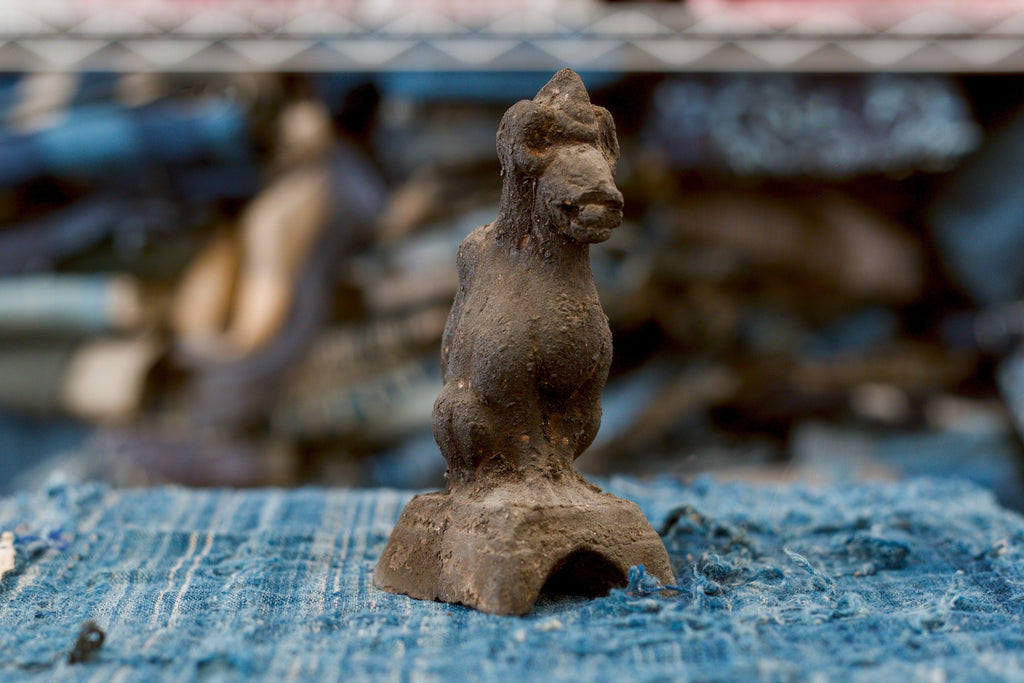 An antique Japanese clay Inari fox on a low pedestal, photographed at a three-quarter angle with its pointed muzzle facing right. The dark, grainy surface covers its tall neck, rounded chest, and seated haunches, while an arched opening is visible in the pedestal above frayed blue cloth.