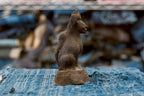 An antique Japanese terracotta Inari fox shown in side profile, facing right. Its projecting muzzle, pointed ears, rounded haunch, and thick tail along the back stand out against the rough, dark-brown surface. The seated figure rests on a square pedestal over worn blue fabric.