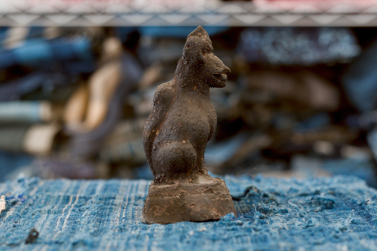 An antique Japanese terracotta Inari fox shown in side profile, facing right. Its projecting muzzle, pointed ears, rounded haunch, and thick tail along the back stand out against the rough, dark-brown surface. The seated figure rests on a square pedestal over worn blue fabric.