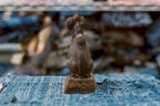 Back view of a small antique Japanese clay Inari fox, showing its tall ears, slim neck, broad seated haunches, and thick tail descending toward the base. Its uneven charcoal-brown surface has scattered warm reddish patches, and the square pedestal rests on frayed blue fabric.