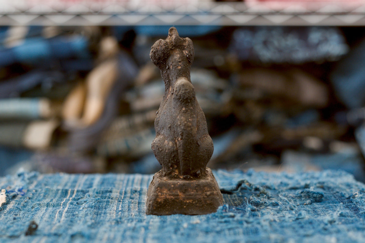 Back view of a small antique Japanese clay Inari fox, showing its tall ears, slim neck, broad seated haunches, and thick tail descending toward the base. Its uneven charcoal-brown surface has scattered warm reddish patches, and the square pedestal rests on frayed blue fabric.