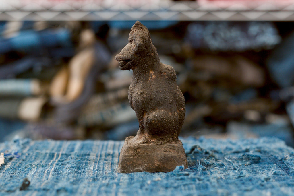 An antique Japanese clay Inari fox in side profile, facing left, with a prominent pointed muzzle, long upright neck, rounded chest, and full haunch. A raised tail follows the back of the dark, heavily textured figure, which sits on a low pedestal over faded blue woven cloth.