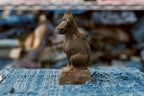 An antique Japanese clay Inari fox in side profile, facing left, with a prominent pointed muzzle, long upright neck, rounded chest, and full haunch. A raised tail follows the back of the dark, heavily textured figure, which sits on a low pedestal over faded blue woven cloth.