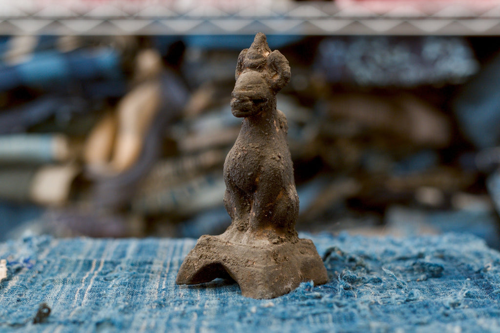 An antique Japanese terracotta Inari fox viewed at a three-quarter angle, its muzzle pointing left above a long neck and rounded chest. The worn dark-brown clay figure sits on a broad pedestal with a curved opening beneath the front, surrounded by frayed blue textile.