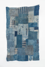 Full-length view of an antique Japanese boro textile hanging vertically, made from faded indigo cotton patches in solids, stripes, and checks, with irregular worn edges.