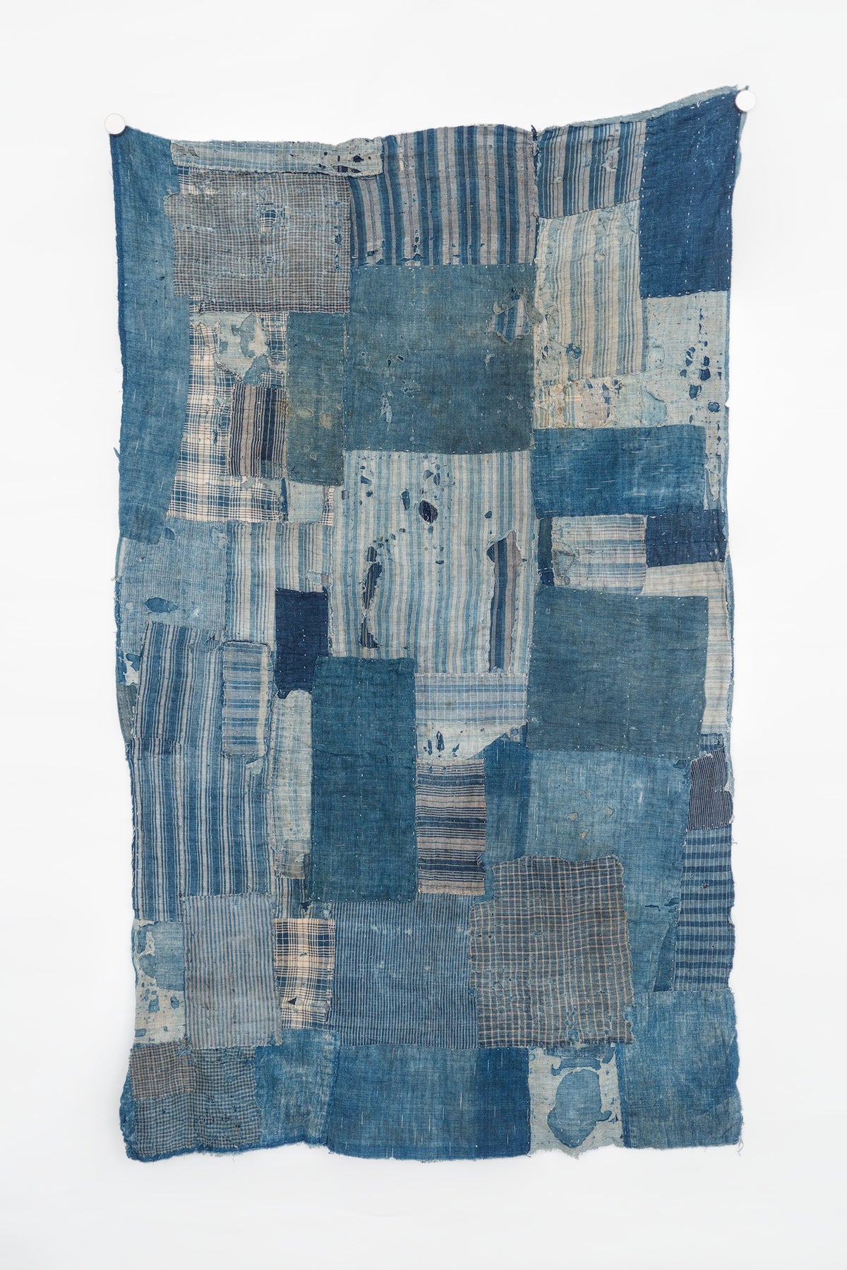 Full-length view of an antique Japanese boro textile hanging vertically, made from faded indigo cotton patches in solids, stripes, and checks, with irregular worn edges.