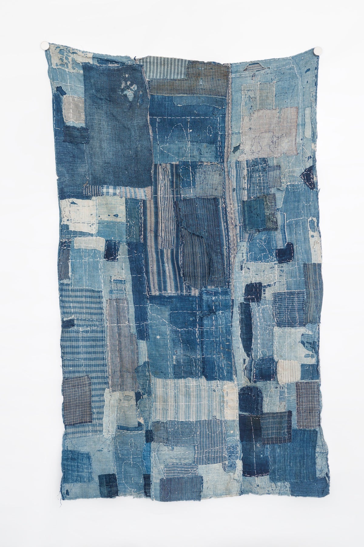 Full-length view of an antique Japanese boro textile hanging vertically, with layered indigo cotton patches, pale blue repairs, stripes, checks, exposed seams, and dense white sashiko stitching.